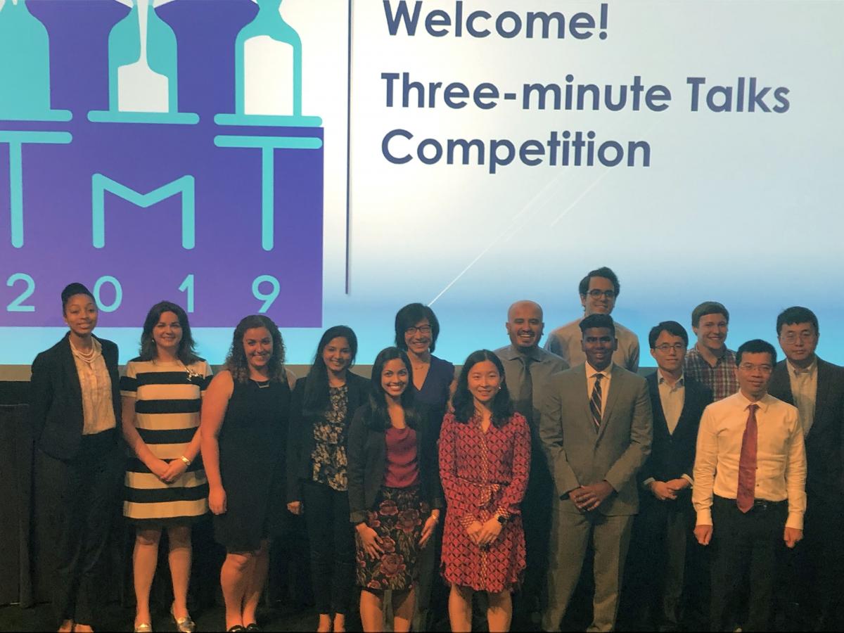 A group of people stands in front of a screen that reads "Welcome! Three-minute Talks Competition."