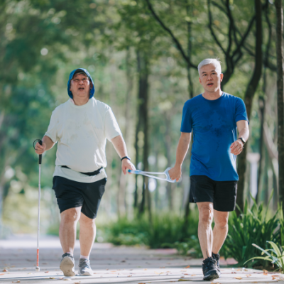  An adult with a visual impairment and a friend walk through a park on a nice day. Each friend holds an end of a tether.