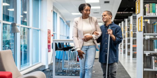 An adult with a visual impairment walking arm in arm with a loved one through a library. 