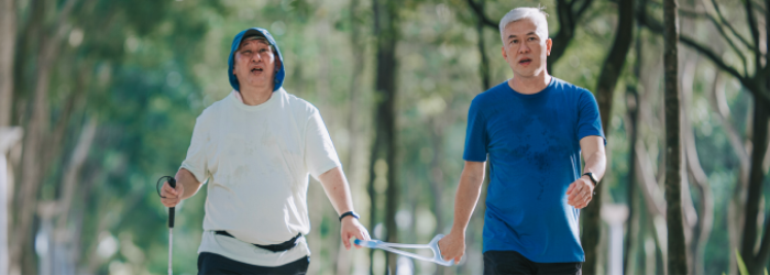 An adult with a visual impairment and a friend taking a brisk walk through a park on a nice day. Each friend is holding on to 1 end of a tether. 