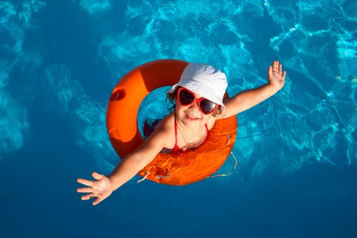 An image of a female preschool child floating in a pool with an orange floaty, a white sunhat, and red heart shaped sunglasses.