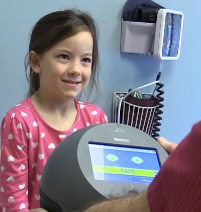 girl in eye provider's office gazes at provider holding the pediatric vision screener. Result shows a "pass" result, meaning she does not need a referral for amblyopia.